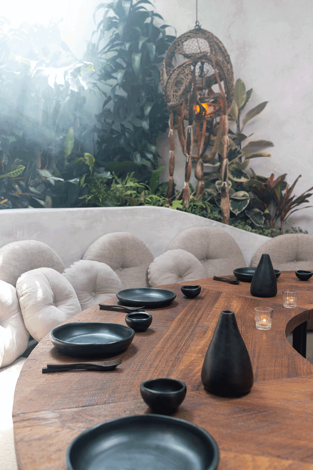 A set wooden table with black ceramic earthenware in front of a linen-colored banquette and tropical plants in the private dining room at meteora